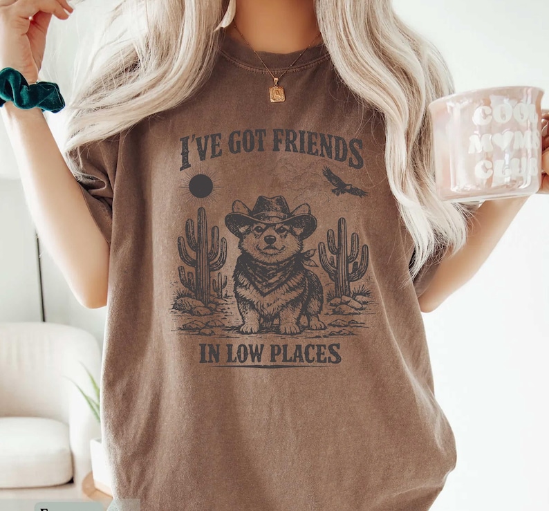 May include: Brown t-shirt with a graphic of a corgi wearing a cowboy hat and bandana, with cacti and the text "I've Got Friends In Low Places". A sun and bird are also in the design. The shirt is a neutral color.