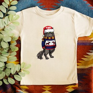 K&ouml;nnte beinhalten: Ein cremefarbenes T-Shirt mit einer schwarzen Katze, die einen roten Weihnachtsmann-Hut und einen blauen Pullover mit Rentieren tr&auml;gt. Der Text "MY MODE" steht auf dem Hut.