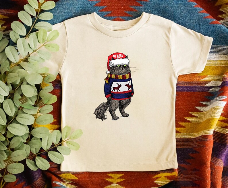 K&ouml;nnte beinhalten: Ein cremefarbenes T-Shirt mit einer schwarzen Katze, die einen roten Weihnachtsmann-Hut und einen blauen Pullover mit Rentieren tr&auml;gt. Der Text "MY MODE" steht auf dem Hut.