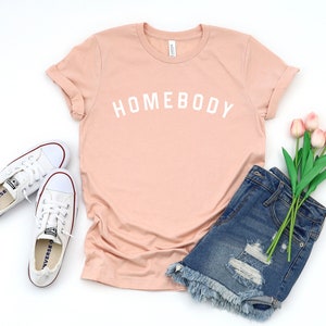 Puede incluir: Una camiseta rosa claro con el texto "HOMEBODY" en letras blancas. La camiseta se combina con pantalones cortos de mezclilla, zapatillas blancas y un ramo de tulipanes rosas.