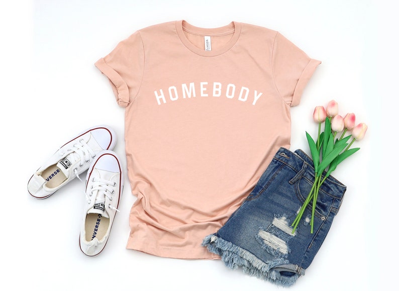 Puede incluir: Una camiseta rosa claro con el texto "HOMEBODY" en letras blancas. La camiseta se combina con pantalones cortos de mezclilla, zapatillas blancas y un ramo de tulipanes rosas.