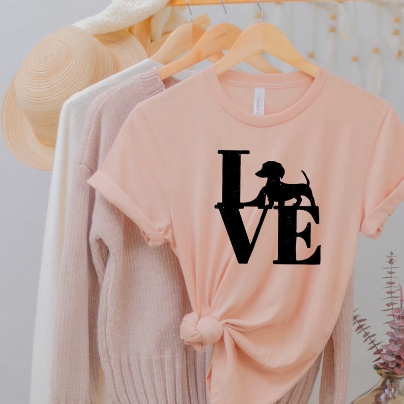 以下が含まれることがあります： ピーチ色のTシャツに、黒いシルエットのダックスフントが「LOVE」の文字の中に描かれています。