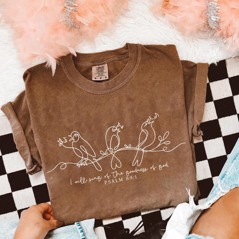 May include: Brown Comfort Colors t-shirt featuring a white line drawing of three birds perched on a branch. The text "I will sing of the goodness of god PSALM 89:1" is printed below the birds. The shirt has a crew neck and short sleeves.