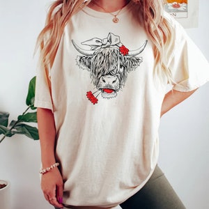 May include: A woman wearing a cream colored t-shirt with a black and white illustration of a Highland cow wearing a red flower headband and holding a red flower in its mouth. The woman is also wearing green biker shorts.