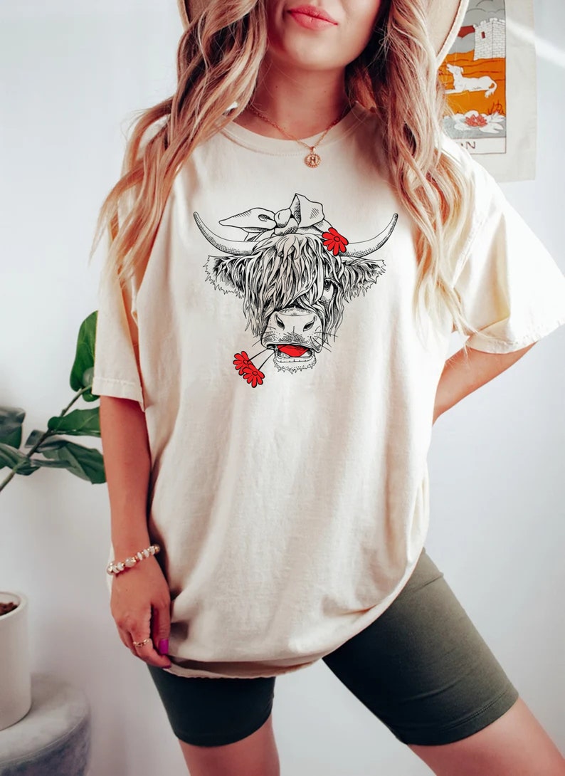 May include: A woman wearing a cream colored t-shirt with a black and white illustration of a Highland cow wearing a red flower headband and holding a red flower in its mouth. The woman is also wearing green biker shorts.