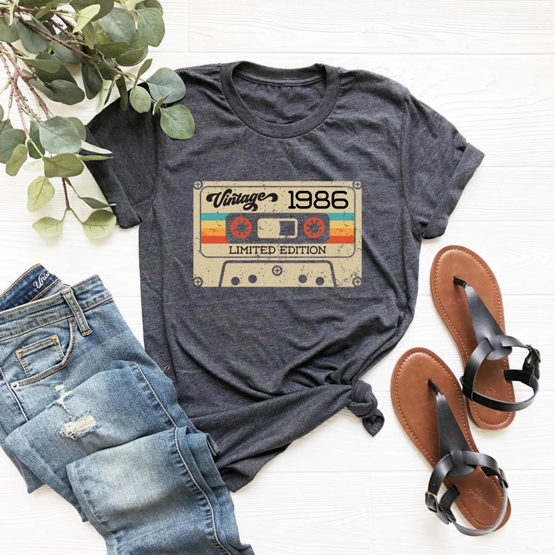 May include: A dark grey t-shirt featuring a vintage cassette tape graphic. The tape has a rainbow design and the text "Vintage 1986 Limited Edition".