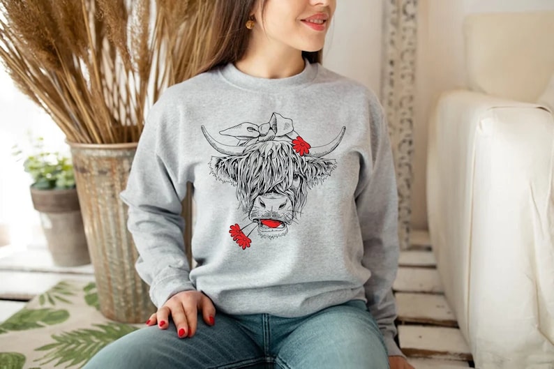 May include: A gray sweatshirt with a black and white illustration of a Highland cow wearing a red bandana and holding red flowers in its mouth.