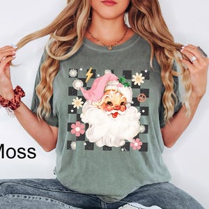 May include: Moss green t-shirt featuring a retro Santa Claus graphic. The Santa Claus design includes a pink hat, white beard, and a checkered background with flowers and peace signs. The word "Moss" is printed on the bottom left.