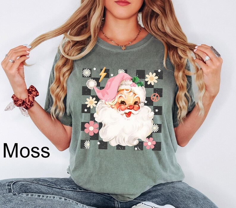 May include: Moss green t-shirt featuring a retro Santa Claus graphic. The Santa Claus design includes a pink hat, white beard, and a checkered background with flowers and peace signs. The word "Moss" is printed on the bottom left.
