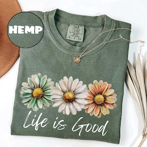 May include: A sage green t-shirt with a daisy graphic and the phrase "Life is Good" in white script. The shirt features a crew neck and short sleeves. A gold necklace with a pendant is draped on the shirt. The word "HEMP" is in a circle.