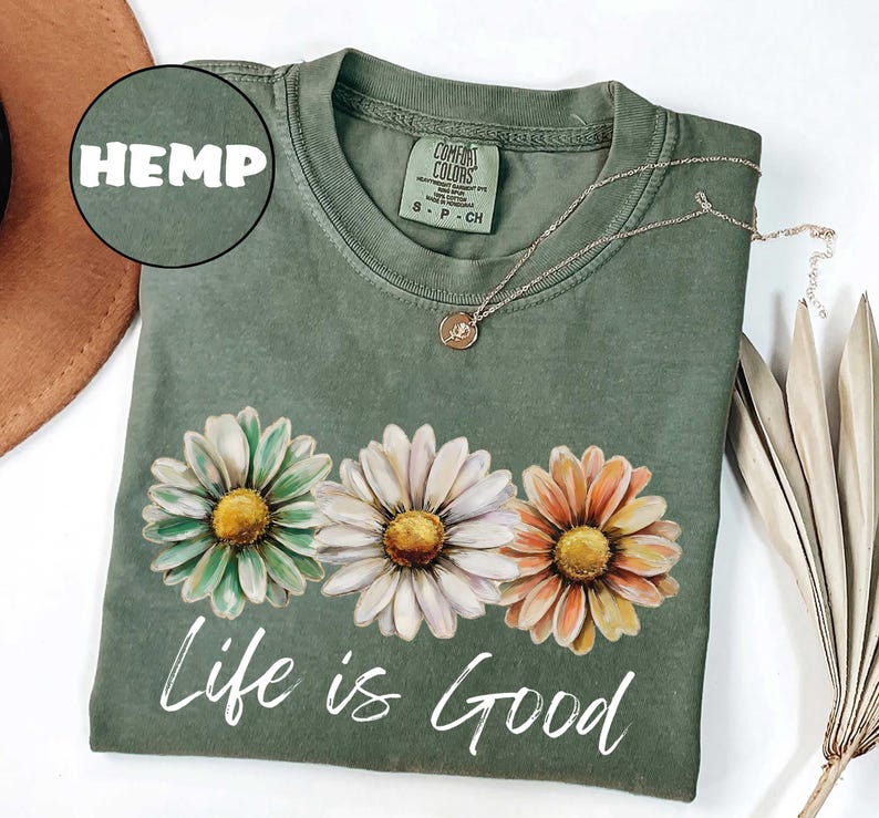 May include: A sage green t-shirt with a daisy graphic and the phrase "Life is Good" in white script. The shirt features a crew neck and short sleeves. A gold necklace with a pendant is draped on the shirt. The word "HEMP" is in a circle.