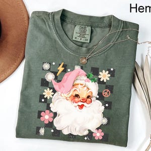 May include: Hemp-colored t-shirt featuring a retro Santa Claus graphic with a pink hat, white beard, and floral accents. The design includes a checkerboard background, a peace sign, and a lightning bolt. A silver necklace rests on the shirt.