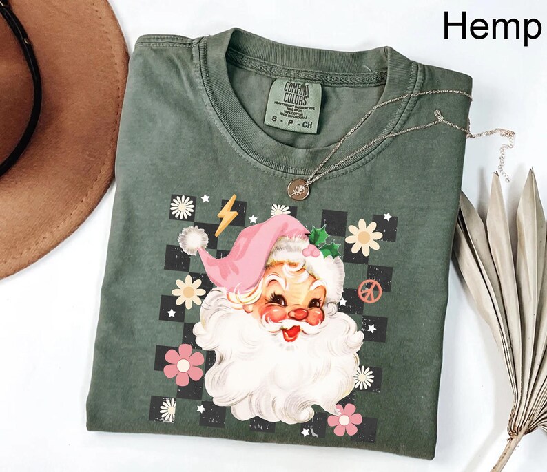 May include: Hemp-colored t-shirt featuring a retro Santa Claus graphic with a pink hat, white beard, and floral accents. The design includes a checkerboard background, a peace sign, and a lightning bolt. A silver necklace rests on the shirt.