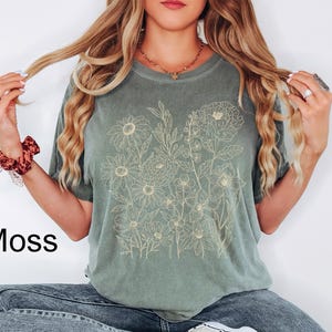 K&ouml;nnte beinhalten: Ein moosgr&uuml;nes T-Shirt mit einem hellbeigen Blumendesign. Das Wort "Moss" ist links unten in Schwarz aufgedruckt. Das Shirt ist ein l&auml;ssiger Stil, geeignet f&uuml;r den Alltag.