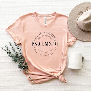 May include: A light pink t-shirt with a black circle graphic that says "God is my refuge and my fortress, Psalms 91, I will find shelter under his wings".