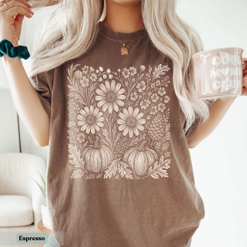 May include: Espresso-colored t-shirt featuring a detailed floral and pumpkin design. The graphic includes large daisies, smaller flowers, and two pumpkins. The shirt is a neutral color, perfect for fall.
