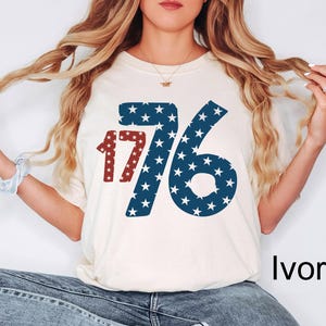 Puede incluir: Una camiseta blanca con un gr&aacute;fico rojo y azul del n&uacute;mero 1776 con estrellas. La camiseta est&aacute; etiquetada como "Marfil".