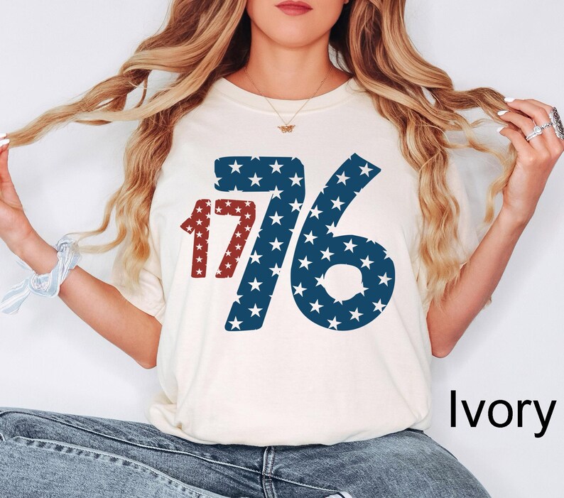 Puede incluir: Una camiseta blanca con un gr&aacute;fico rojo y azul del n&uacute;mero 1776 con estrellas. La camiseta est&aacute; etiquetada como "Marfil".