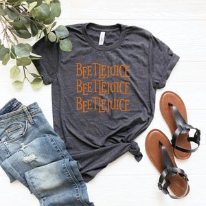 May include: A dark gray t-shirt with the words "Beetlejuice" repeated three times in orange lettering. The shirt is laid out on a white surface with a pair of brown sandals and blue jeans.