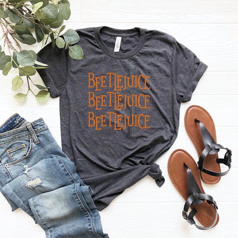 May include: A dark gray t-shirt with the words "Beetlejuice" repeated three times in orange lettering. The shirt is laid out on a white surface with a pair of brown sandals and blue jeans.