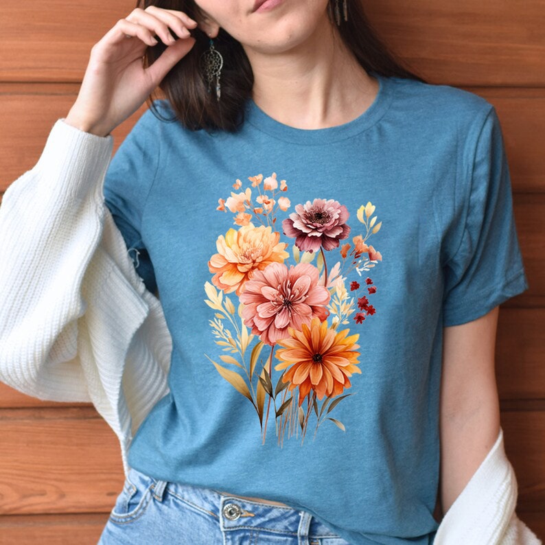 May include: A teal blue t-shirt with a floral print featuring a bouquet of orange, pink, and brown flowers. The flowers are watercolor style and have a vintage aesthetic.