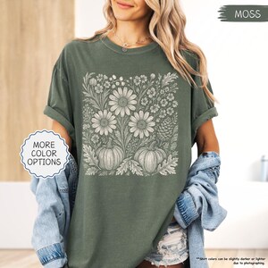 May include: A moss green t-shirt featuring a floral and pumpkin design in white. The design includes large daisies, smaller flowers, and two pumpkins. The words "MORE COLOR OPTIONS" are in a white circle. The word "MOSS" is in a green rectangle.