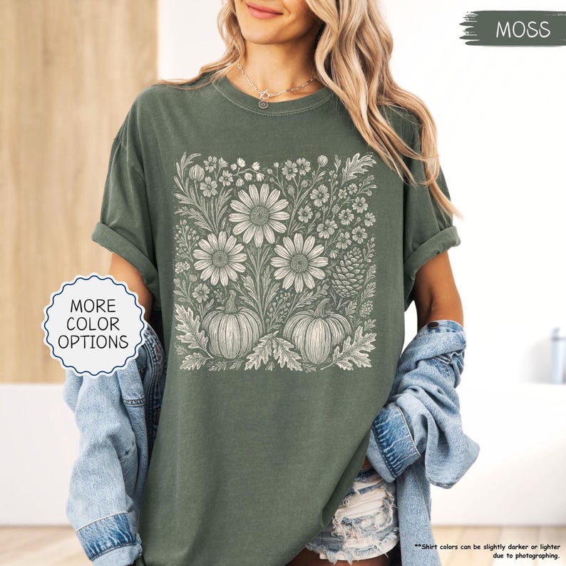 May include: A moss green t-shirt featuring a floral and pumpkin design in white. The design includes large daisies, smaller flowers, and two pumpkins. The words "MORE COLOR OPTIONS" are in a white circle. The word "MOSS" is in a green rectangle.