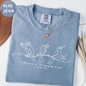 May include: A blue jean Comfort Colors t-shirt featuring a white line drawing of three birds perched on a branch. The text "I will sing of the goodness of god, Psalm 89:1" is printed below. A gold necklace is placed on the shirt.