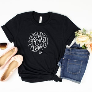 May include: Black t-shirt with white text that reads "Stay Groovy" in a retro font. The shirt is styled with denim shorts, pink flats, and sunglasses.