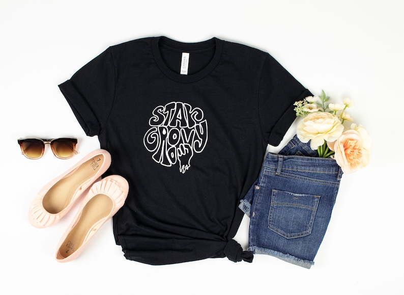 May include: Black t-shirt with white text that reads "Stay Groovy" in a retro font. The shirt is styled with denim shorts, pink flats, and sunglasses.