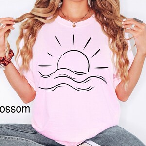 Puede incluir: Camiseta rosa claro con un dise&ntilde;o negro de un sol y olas. La palabra "Blossom" est&aacute; impresa en negro en la parte inferior izquierda. La camiseta es de material suave y es adecuada para uso casual.
