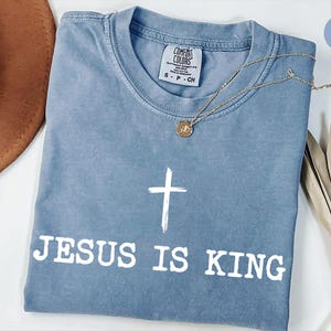 K&ouml;nnte beinhalten: Blaues Jeans-T-Shirt mit dem wei&szlig;en Text &bdquo;JESUS IS KING&ldquo; und einem wei&szlig;en Kreuz. Eine goldene Halskette mit Anh&auml;nger liegt auf dem T-Shirt. Das T-Shirt ist auf einer wei&szlig;en Oberfl&auml;che gefaltet. Comfort Colors Marke.