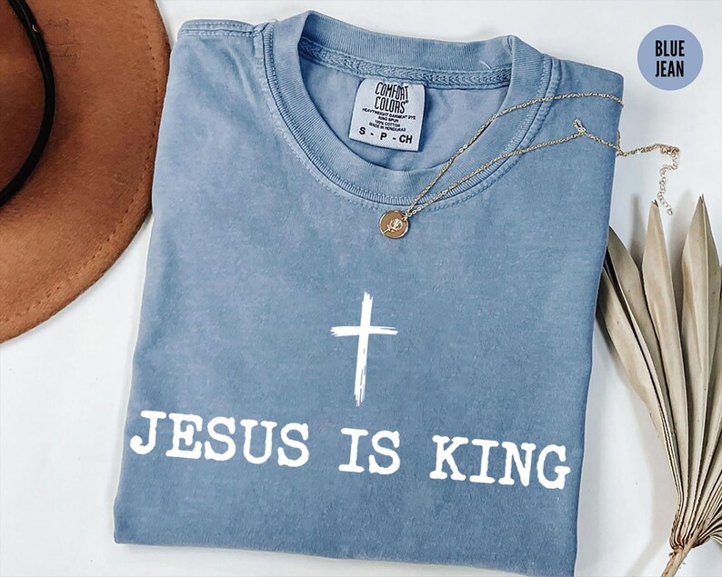 K&ouml;nnte beinhalten: Blaues Jeans-T-Shirt mit dem wei&szlig;en Text &bdquo;JESUS IS KING&ldquo; und einem wei&szlig;en Kreuz. Eine goldene Halskette mit Anh&auml;nger liegt auf dem T-Shirt. Das T-Shirt ist auf einer wei&szlig;en Oberfl&auml;che gefaltet. Comfort Colors Marke.