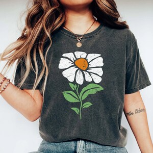 May include: A dark gray t-shirt with a white daisy flower graphic. The flower has a yellow center and green leaves. The shirt has a distressed look.