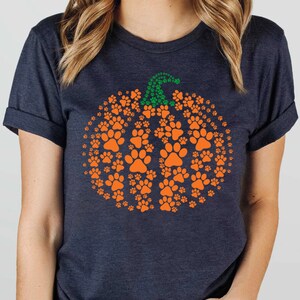 Peut inclure: T-shirt bleu marine fonc&eacute; avec un motif de citrouille orange compos&eacute; d'empreintes de pattes. La citrouille a une tige verte faite de petites empreintes de pattes. Un design amusant et festif pour les amoureux des animaux.