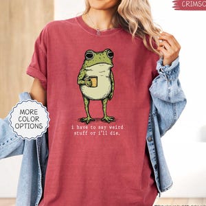 May include: Crimson-colored t-shirt featuring a green frog holding a mug. The frog illustration has a vintage style. Text on the shirt reads, "I have to say weird stuff or I'll die." A "More Color Options" button is visible.