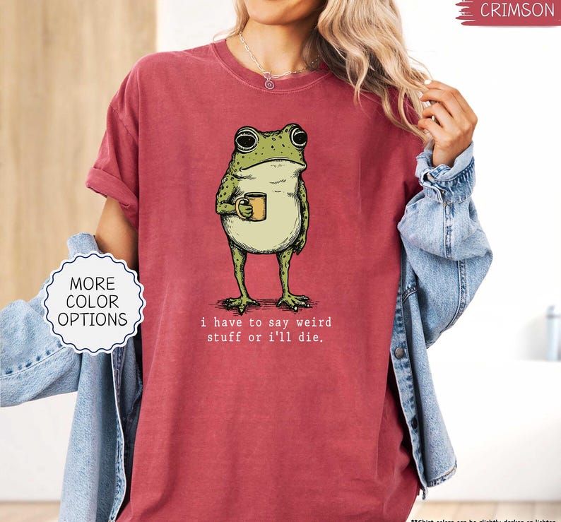 May include: Crimson-colored t-shirt featuring a green frog holding a mug. The frog illustration has a vintage style. The text "I have to say weird stuff or I'll die" is printed below the frog. The shirt has a relaxed fit and rolled sleeves.