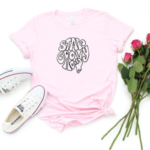 May include: A pink t-shirt with the text "Stay Groovy" in a retro font. The shirt is laid flat on a white background with a pair of white sneakers and a bouquet of pink roses.