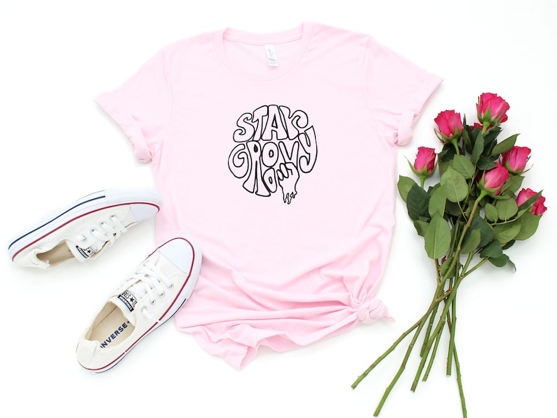 May include: A pink t-shirt with the text "Stay Groovy" in a retro font. The shirt is laid flat on a white background with a pair of white sneakers and a bouquet of pink roses.
