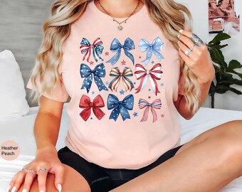 May include: Heather peach-colored t-shirt featuring a grid of watercolor bows in red, white, and blue. The bows have star and stripe patterns, perfect for patriotic celebrations. The shirt is a short-sleeved crew neck.