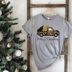 Op de afbeelding: Een grijs T-shirt met een pick-up truck in luipaardprint die een kerstboom vervoert. De truck heeft gouden accenten en de tekst "Merry Christmas" is onder de truck gedrukt.