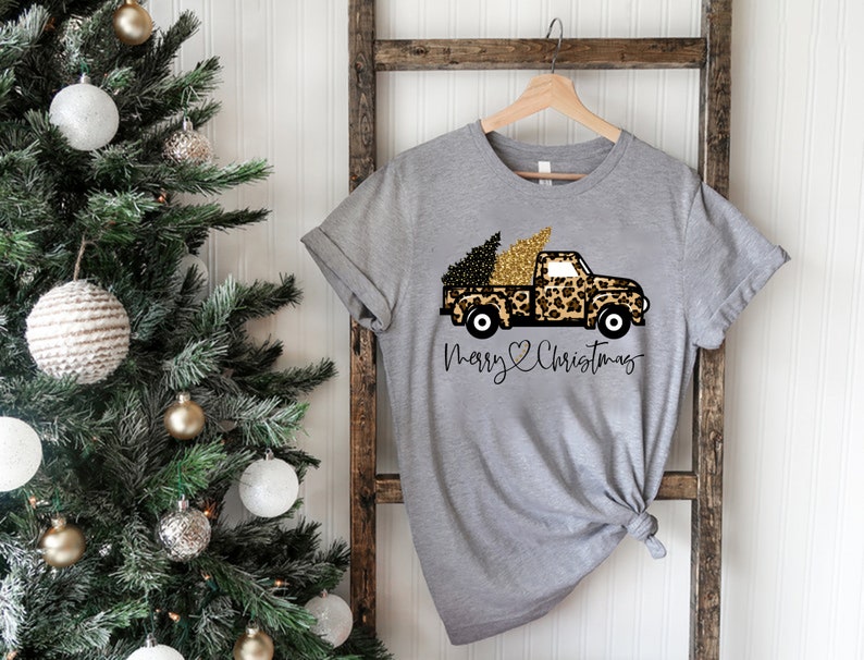 Op de afbeelding: Een grijs T-shirt met een pick-up truck in luipaardprint die een kerstboom vervoert. De truck heeft gouden accenten en de tekst "Merry Christmas" is onder de truck gedrukt.