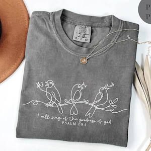 May include: A grey t-shirt featuring a white line drawing of three birds perched on a branch. The text "I will sing of the goodness of god PSALM 89:1" is printed below. A silver necklace with a small pendant rests on the shirt.