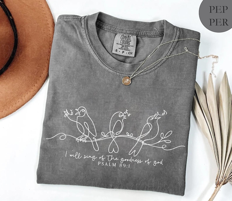 May include: A grey t-shirt featuring a white line drawing of three birds perched on a branch. The text "I will sing of the goodness of god PSALM 89:1" is printed below. A silver necklace with a small pendant rests on the shirt.