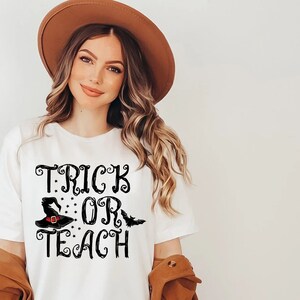 Puede incluir: Camiseta blanca con la frase "TRICK OR TEACH" en letras negras. El dise&ntilde;o incluye un sombrero de bruja y un murci&eacute;lago. Ideal para Halloween o para profesores.
