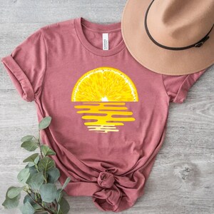 Pu&ograve; includere: Una maglietta rosa con una grafica di una fetta di limone su un tramonto giallo.