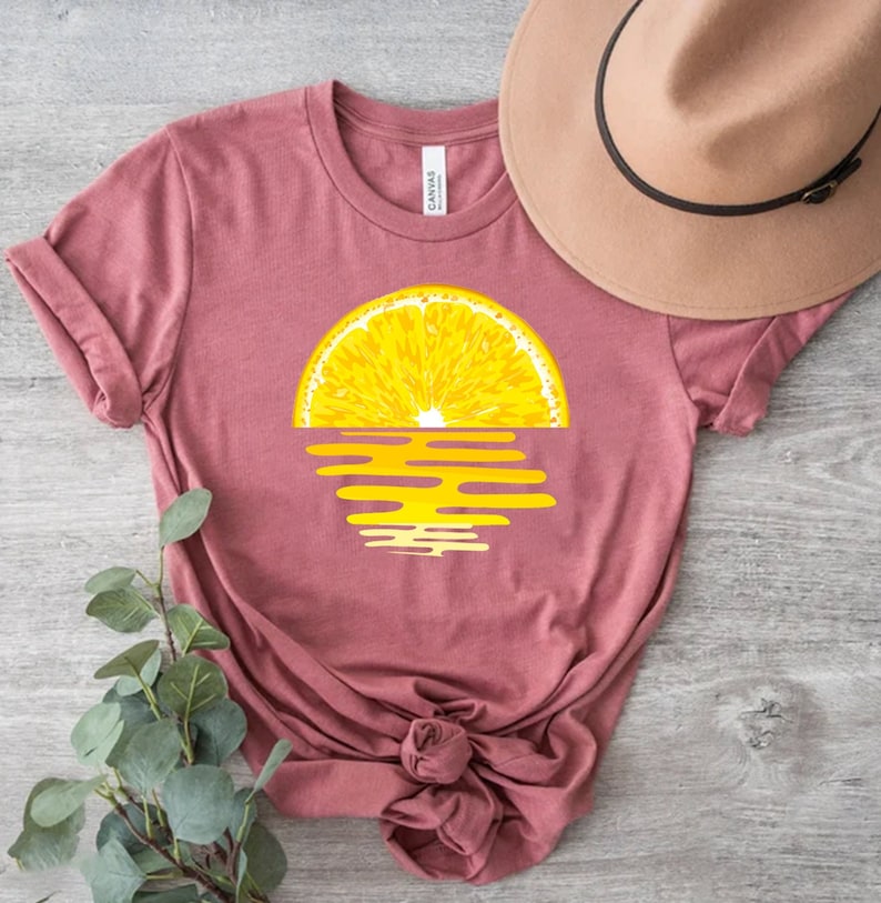Pu&ograve; includere: Una maglietta rosa con una grafica di una fetta di limone su un tramonto giallo.