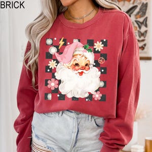 May include: Brick-colored sweatshirt featuring a retro Santa Claus graphic. The Santa design includes a pink hat, white beard, and is surrounded by flowers and a checkered pattern. The word "BRICK" is printed at the top.