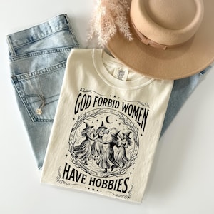 K&ouml;nnte beinhalten: Cremefarbenes T-Shirt mit dem Text "GOD FORBID WOMEN HAVE HOBBIES" in Schwarz. Das Shirt zeigt eine Grafik von vier Hexen in einem Kreis. Das Shirt wird mit hellblauen Jeans, einem beigen Hut und einer goldenen Halskette kombiniert.