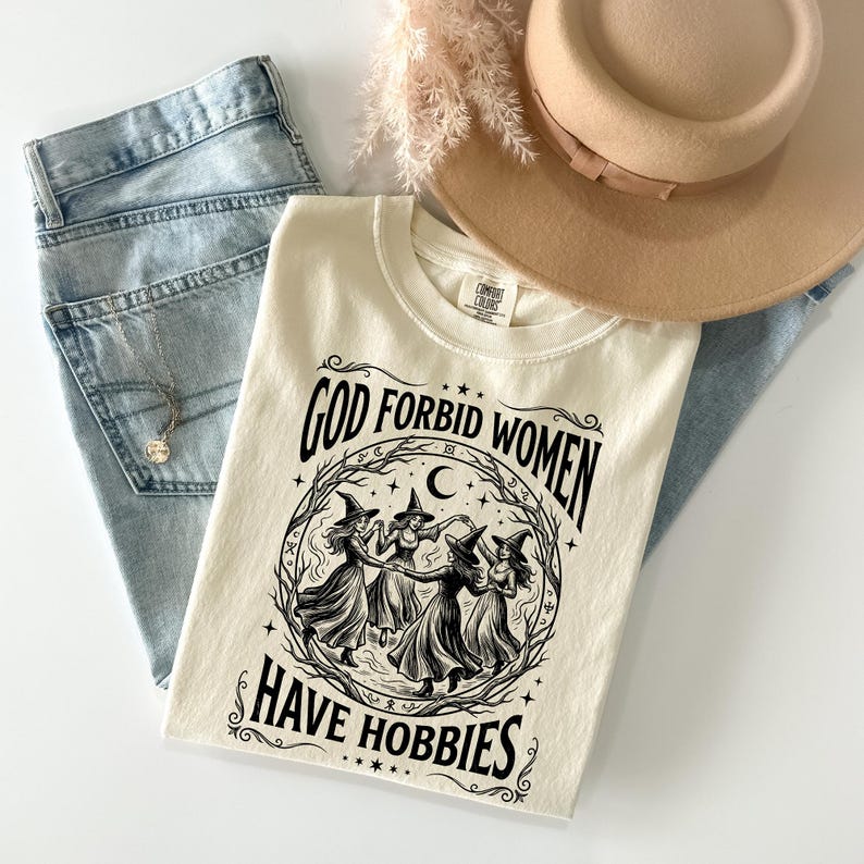 K&ouml;nnte beinhalten: Cremefarbenes T-Shirt mit dem Text "GOD FORBID WOMEN HAVE HOBBIES" in Schwarz. Das Shirt zeigt eine Grafik von vier Hexen in einem Kreis. Das Shirt wird mit hellblauen Jeans, einem beigen Hut und einer goldenen Halskette kombiniert.